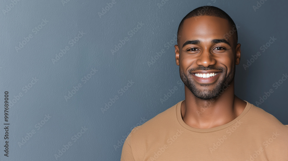 Obraz premium A confident african american man with a bright smile stands against a gray wall, exuding positivity and approachability with a casual tan shirt and relaxed demeanor, copy space