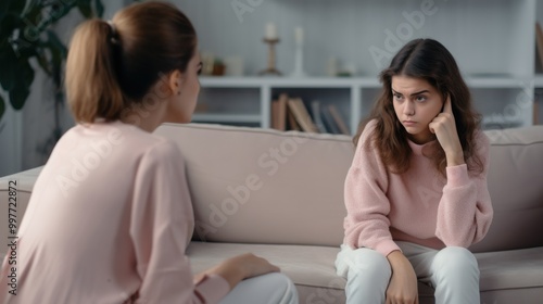 A concerned mother in a pink sweater tries to communicate with her visibly upset teenage daughter, who is seated on a sofa and looking away.