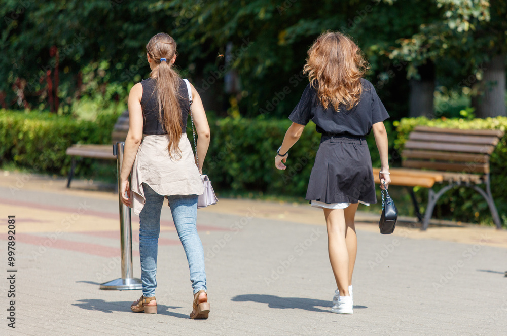 Two women walking down a sidewalk, one of them carrying a purse Two women walking down a sidewalk, one of them carrying a purse