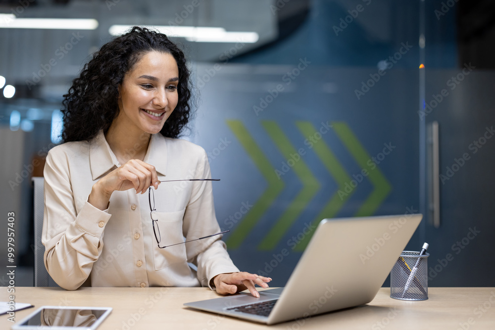 Businesswoman smiling while working on laptop, holding glasses. Professional setting suggests office work, focus, and success. Mixed ethnicity woman engaged in business tasks, showcasing motivation Businesswoman smiling while working on laptop, holding glasses. Professional setting suggests office work, focus, and success. Mixed ethnicity woman engaged in business tasks, showcasing motivation