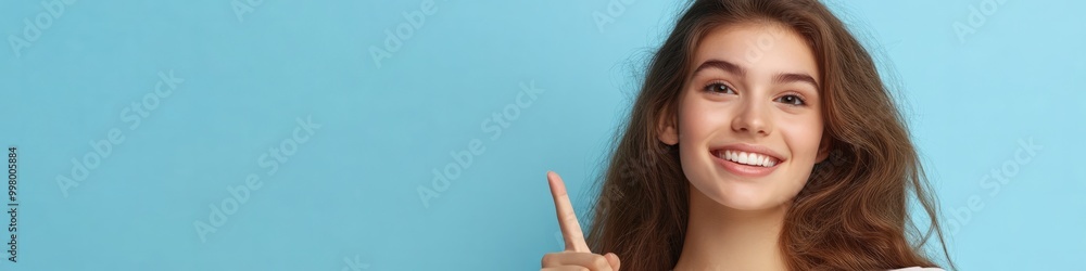 Confident and Poised: A young woman with a bright smile points upward, radiating positivity and enthusiasm against a vibrant blue backdrop. Her gesture suggests a new direction, a brilliant idea. Confident and Poised: A young woman with a bright smile points upward, radiating positivity and enthusiasm against a vibrant blue backdrop. Her gesture suggests a new direction, a brilliant idea.