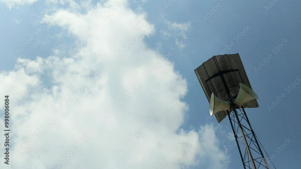 A tall communication tower with dual loudspeakers mounted under a metal roof, set against a bright blue sky with light clouds. The contrast between the man-made structure and natural sky. A tall communication tower with dual loudspeakers mounted under a metal roof, set against a bright blue sky with light clouds. The contrast between the man-made structure and natural sky.