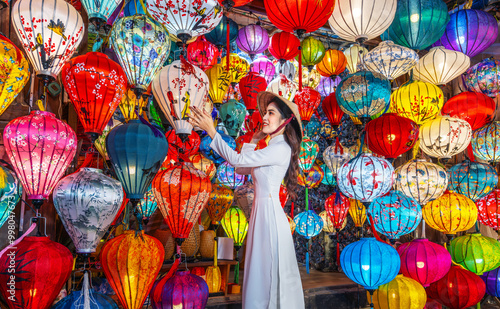 Asian woman wearing Ao Dai Vietnamese dress with colorful lanterns, traveler sightseeing at Hoi An ancient town in central Vietnam.