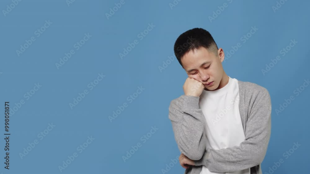 Young asian guy in casual clothes falls asleep standing, resting his head on his hand and then suddenly wakes up with a tired face on a blue background in the studio Young asian guy in casual clothes falls asleep standing, resting his head on his hand and then suddenly wakes up with a tired face on a blue background in the studio