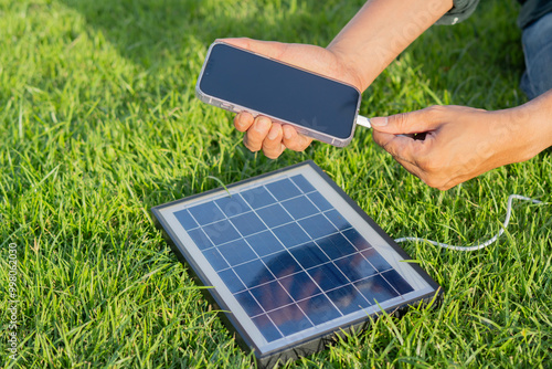 Shot view of man's hnads charging smartphone with solar panel on a lawn in a sunny summer day photo