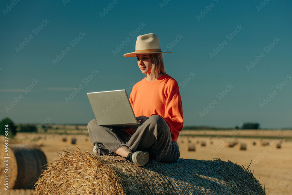 Focused woman wearing hat and using laptop sitting on hay bale in field Focused woman wearing hat and using laptop sitting on hay bale in field