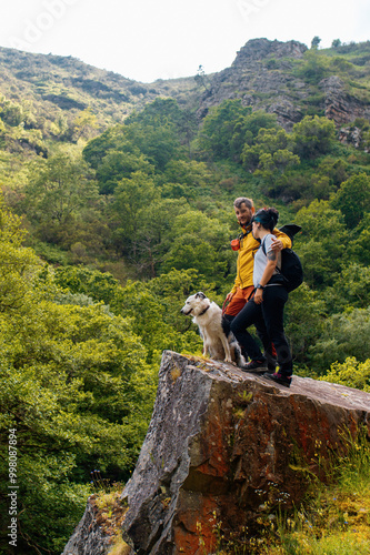 Couple of hikers hugging each other standing on a rock overlooking the lush forest during a day of hiking in the mountains with their bordr collie dog. Hoces del Esva route, Asturias, Spain photo