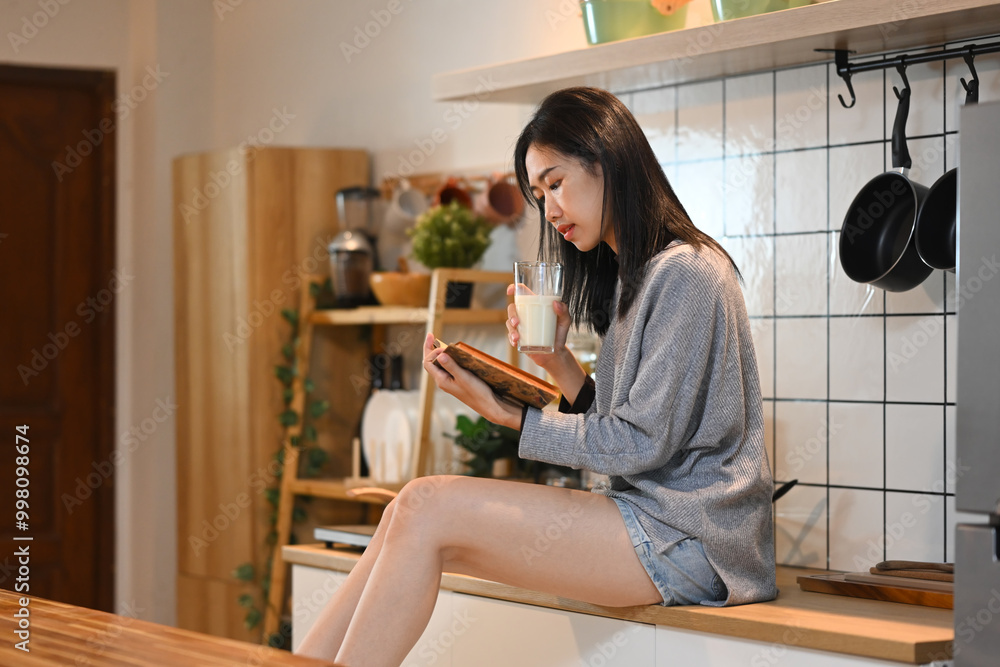 Beautiful young woman sitting on a kitchen countertop, holding a glass of milk and reading a book Beautiful young woman sitting on a kitchen countertop, holding a glass of milk and reading a book