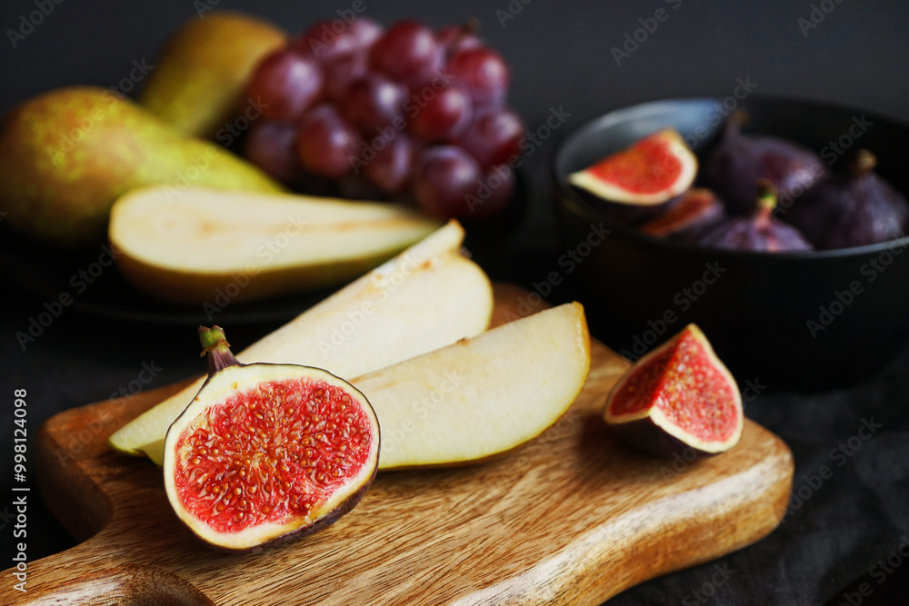 Ripe whole and sliced figs, pears and pink grapes on a wooden board and black plate on a dark background Ripe whole and sliced figs, pears and pink grapes on a wooden board and black plate on a dark background