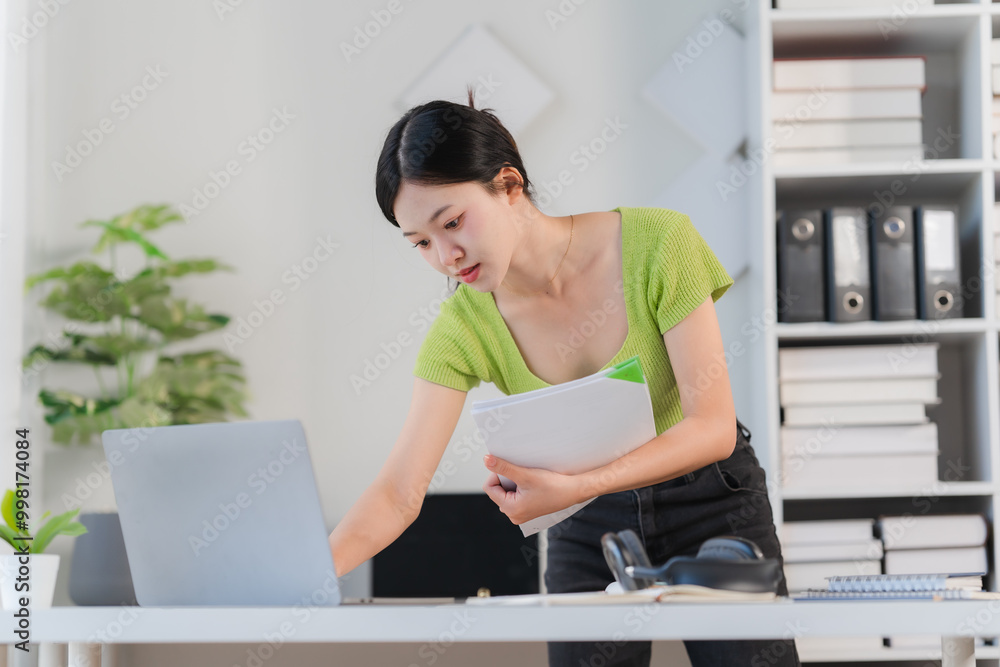 Focused on the Task: A young woman in a green top leans over her laptop, intently reviewing documents in her modern office workspace. Focused on the Task: A young woman in a green top leans over her laptop, intently reviewing documents in her modern office workspace.
