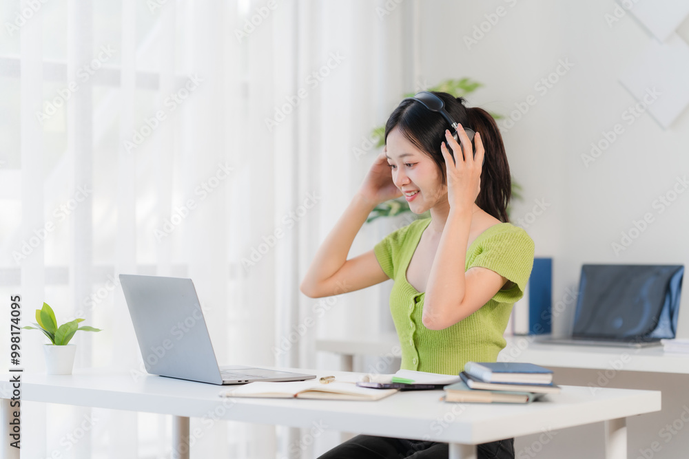Smiling Woman in Online Meeting. A young woman smiles happily while participating in a video conference call on her laptop. She wears headphones and is sitting at a bright, organized desk. Smiling Woman in Online Meeting. A young woman smiles happily while participating in a video conference call on her laptop. She wears headphones and is sitting at a bright, organized desk.