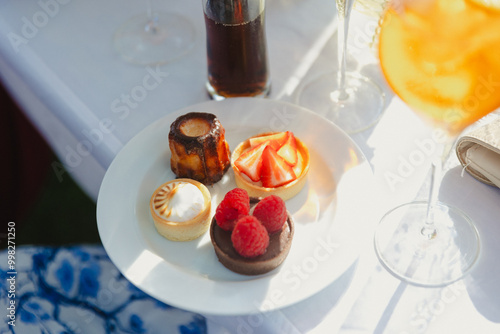 An elegant plate of assorted desserts, including a raspberry-topped tart, strawberry tart, lemon tart, and canelé, served on a white plate at an outdoor event (wedding). photo