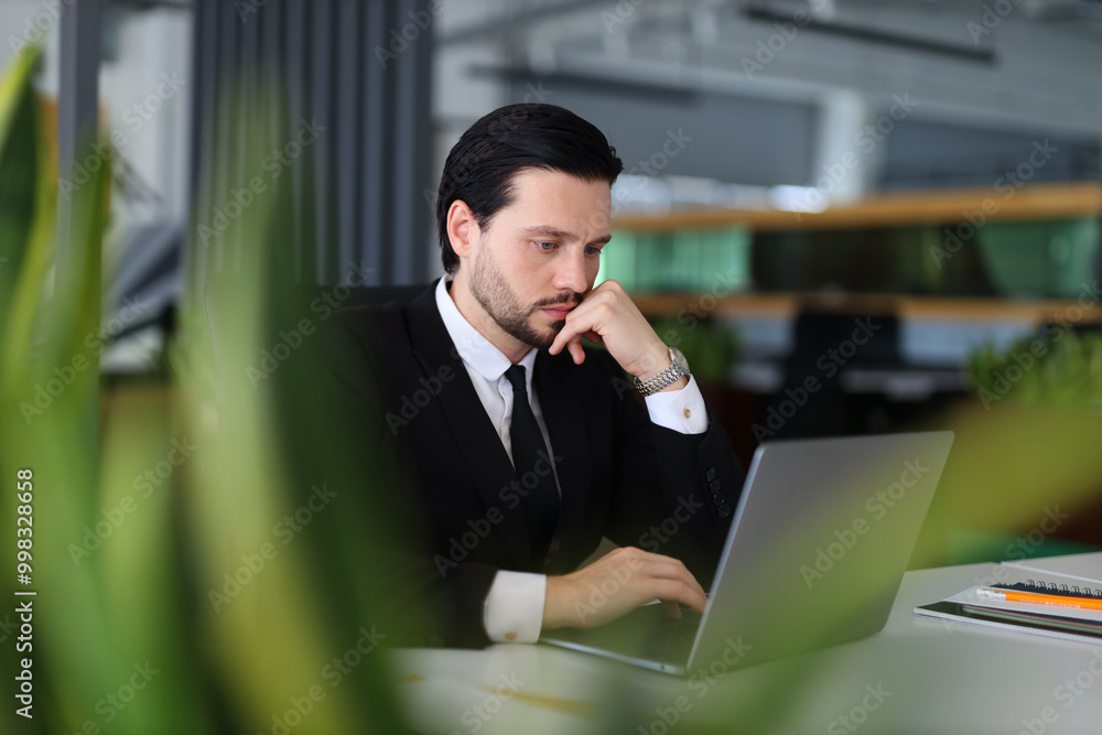 A man in a suit is typing on a laptop computer. He is wearing a watch and has a serious expression on his face. Concept of focus and determination as the man works on his computer