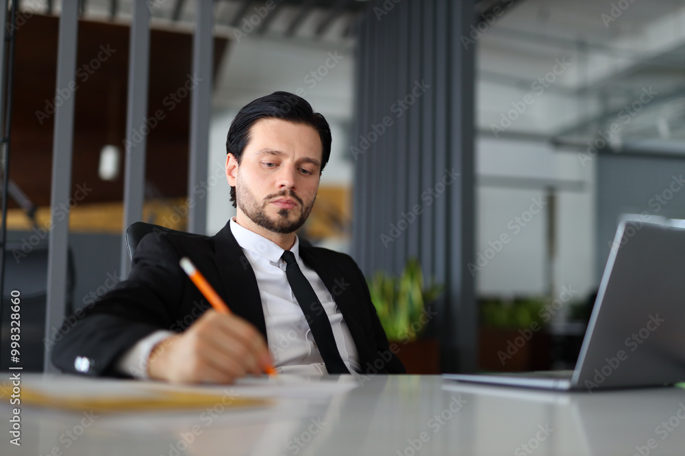 A man in a suit is writing with a pencil on a desk. Concept of professionalism and focus, as the man is likely working on a task or project. The presence of the laptop A man in a suit is writing with a pencil on a desk. Concept of professionalism and focus, as the man is likely working on a task or project. The presence of the laptop