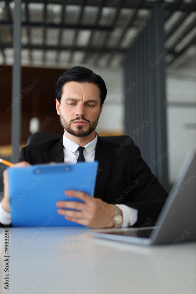 A man in a suit is sitting at a desk with a blue clipboard and a pen. He is focused on the clipboard, possibly taking notes or reviewing some information. Concept of professionalism and concentration A man in a suit is sitting at a desk with a blue clipboard and a pen. He is focused on the clipboard, possibly taking notes or reviewing some information. Concept of professionalism and concentration