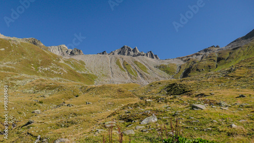 Scenic view of the Novena Pass in the Swiss Alps on a clear summer day, featuring rocky slopes, grassy hills, and a vivid blue sky.