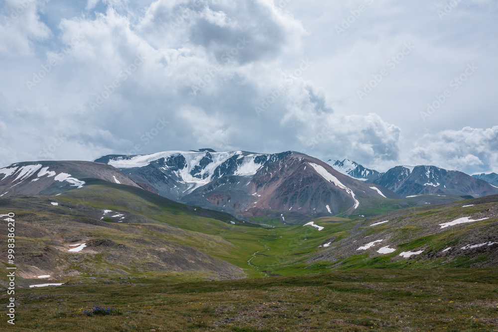 Dramatic view to large snow-capped mountain range under gray rainy cloudy sky. Shadows of clouds on green valley and stony hills. Snowy rocky mountain wall. Changeable weather in big snow mountains. Dramatic view to large snow-capped mountain range under gray rainy cloudy sky. Shadows of clouds on green valley and stony hills. Snowy rocky mountain wall. Changeable weather in big snow mountains.