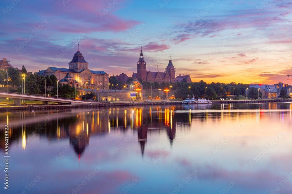 Sunset view of the Szczecin riverfront along the Oder River in Poland, featuring the illuminated Waly Chrobrego buildings and their reflection in the calm water Sunset view of the Szczecin riverfront along the Oder River in Poland, featuring the illuminated Waly Chrobrego buildings and their reflection in the calm water