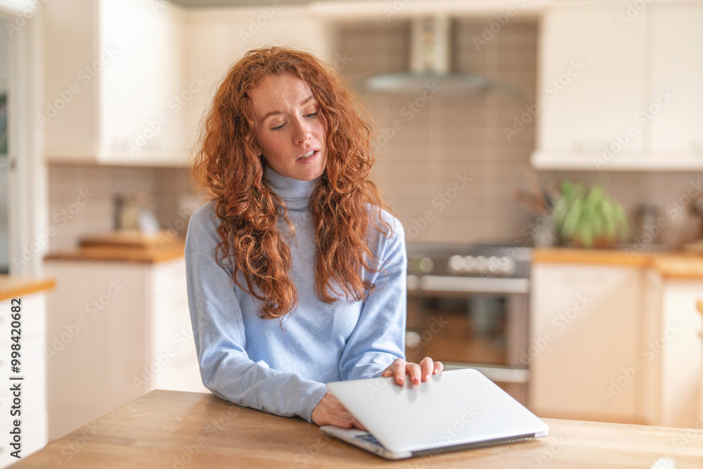 Woman Wearing Blue Sweater is Working on Her Laptop in Bright, Modern Kitchen Woman Wearing Blue Sweater is Working on Her Laptop in Bright, Modern Kitchen