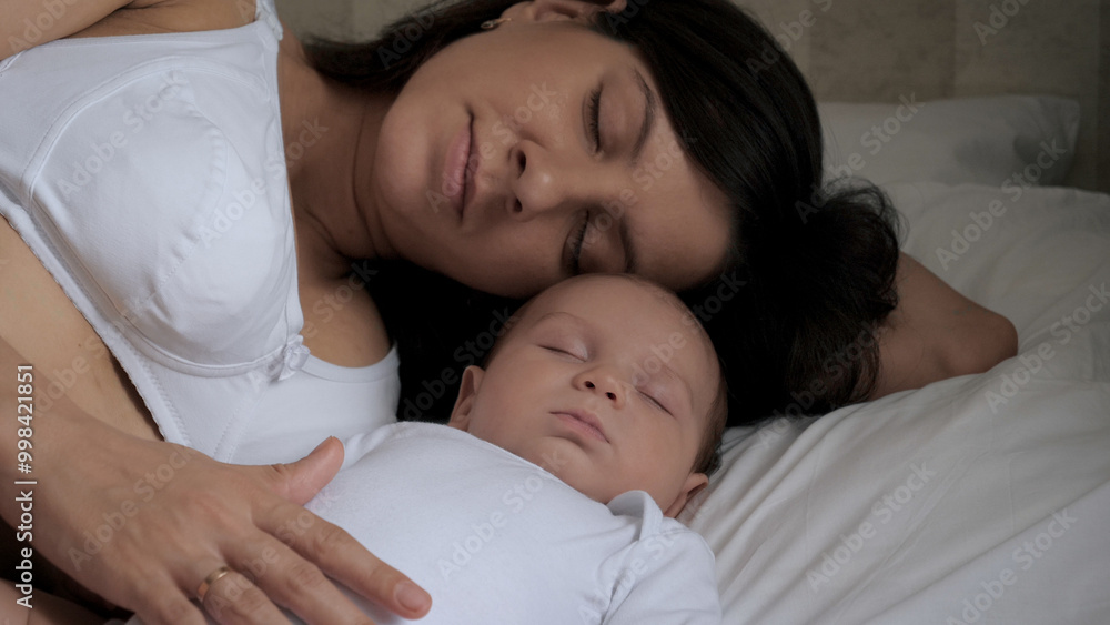 Mom and sleeping newborn baby are lying in bed together. Gently and lovingly strokes baby tummy belly with her hand for daytime sleep in bedroom. Relationship of mother and child. Parental care. Mom and sleeping newborn baby are lying in bed together. Gently and lovingly strokes baby tummy belly with her hand for daytime sleep in bedroom. Relationship of mother and child. Parental care.