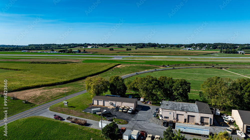 Obraz premium The aerial perspective captures a rural airport with a visible runway, surrounded by fields and greenery. Nearby buildings and vehicles create a sense of activity under a clear blue sky.