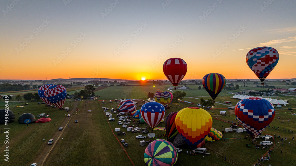 Fototapeta premium A dozen colorful hot air balloons are preparing for launch in a festival field during sunset. Spectators gather to witness the spectacle as the evening sky radiates warm colors.