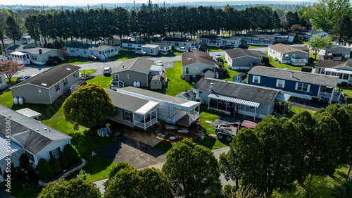 Fototapeta Naklejka Na Ścianę i Meble -  Aerial view shows mobile, manufactured homes lined along a tranquil street, surrounded by well-maintained lawns and trees. The sun casts a warm light, highlighting the neat layout and construction.