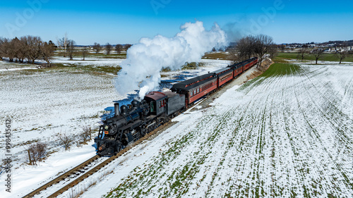 A classic steam locomotive pulls a passenger train along snow-covered tracks in rural territory. The bright sky contrasts with the winter scenery, creating a picturesque travel experience. photo