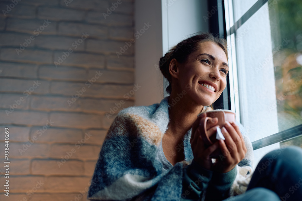 Fototapeta premium Happy woman wrapped in a blanket drinking tea while enjoying by the window at home.