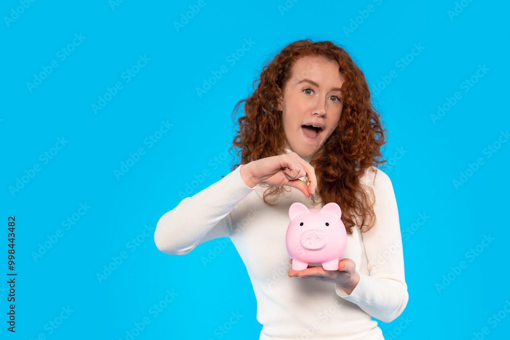 Young Woman Excitedly Saving Money in Pink Piggy Bank on Blue Background Young Woman Excitedly Saving Money in Pink Piggy Bank on Blue Background