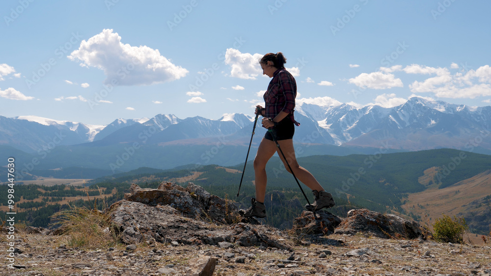Obraz premium Active athletic woman tourist millennial blogger hiking in valley with trekking poles outdoors on sunny day on rocks of hills. Against of snow capped summit of mountains. Summer travel holiday nature