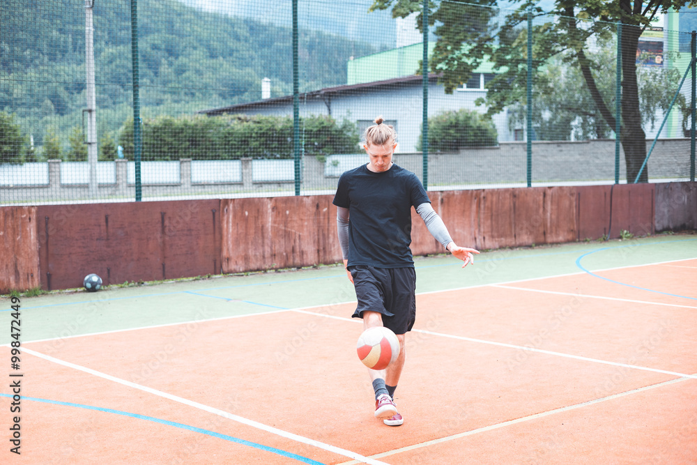 Young man with a man bun juggles a football on a vibrant orange court during a sunny day. Wearing casual sportswear, he focuses on controlling the ball with precision in a natural setting Young man with a man bun juggles a football on a vibrant orange court during a sunny day. Wearing casual sportswear, he focuses on controlling the ball with precision in a natural setting