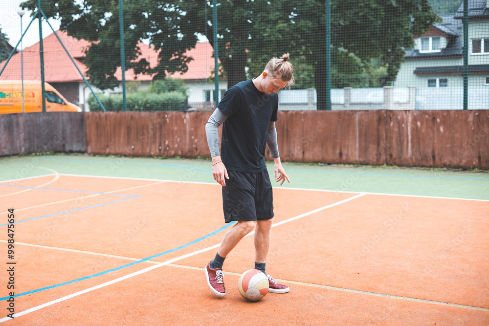 Young man with a man bun juggles a football on a vibrant orange court during a sunny day. Wearing casual sportswear, he focuses on controlling the ball with precision in a natural setting Young man with a man bun juggles a football on a vibrant orange court during a sunny day. Wearing casual sportswear, he focuses on controlling the ball with precision in a natural setting