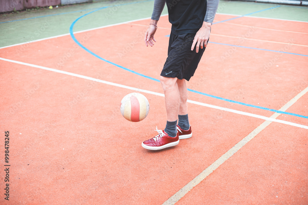 Young man with a man bun juggles a football on a vibrant orange court during a sunny day. Wearing casual sportswear, he focuses on controlling the ball with precision in a natural setting Young man with a man bun juggles a football on a vibrant orange court during a sunny day. Wearing casual sportswear, he focuses on controlling the ball with precision in a natural setting