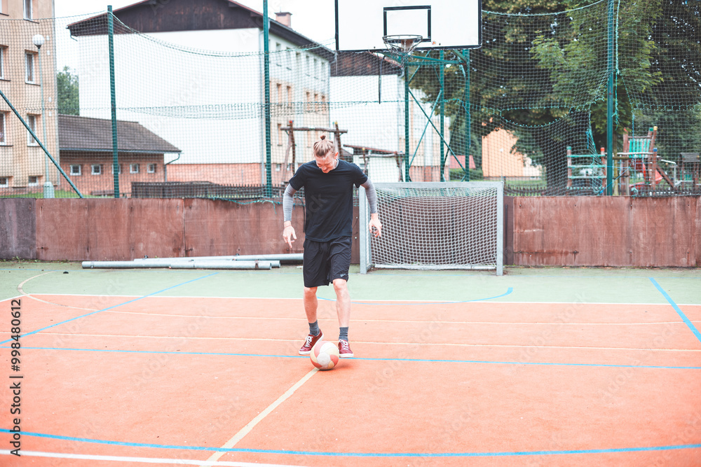 Obraz premium Young man focuses on controlling a football on an outdoor court. He carefully juggles the ball with precision, surrounded by an urban backdrop of buildings and a goal, combining sports and skill