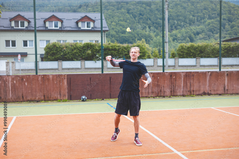 Badminton player makes a powerful serve on an outdoor court, hitting the shuttlecock mid-air. Wearing casual athletic clothing, he is fully focused on the game, with a scenic backdrop of nature Badminton player makes a powerful serve on an outdoor court, hitting the shuttlecock mid-air. Wearing casual athletic clothing, he is fully focused on the game, with a scenic backdrop of nature