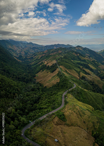 A rural road winds through a serene mountain vista, bordered by lush green forest under a blue sky at dawn in Doi Phuka National Park, Nan Province, Thailand