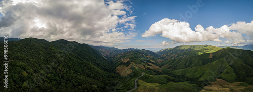 Fotografie A rural road winds through a serene mountain vista, bordered by lush green fores