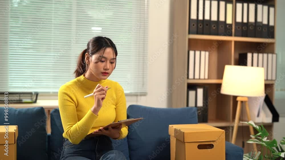 Focused entrepreneur woman managing her business from home, checking inventory list on clipboard. She is sitting on the sofa with cardboard boxes next to her Focused entrepreneur woman managing her business from home, checking inventory list on clipboard. She is sitting on the sofa with cardboard boxes next to her