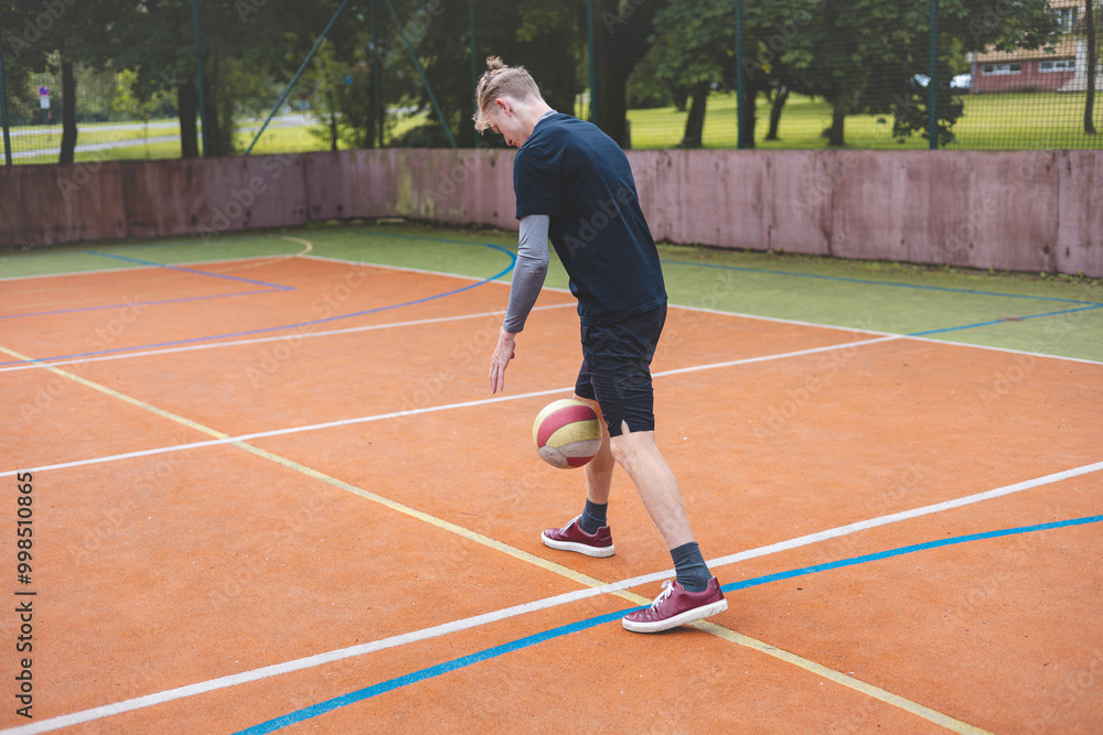 Obraz premium Young man dribbles a basketball on an outdoor court surrounded by nature. Dressed casually in sports attire, he showcases focus and skill during a casual practice session in a vibrant setting