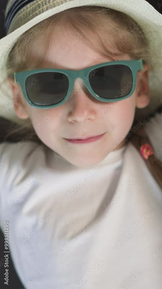 Little girl in straw hat and stylish sunglasses looks in camera lying on sofa. Cute preschooler in white t-shirt enjoys relaxing upper close view Little girl in straw hat and stylish sunglasses looks in camera lying on sofa. Cute preschooler in white t-shirt enjoys relaxing upper close view