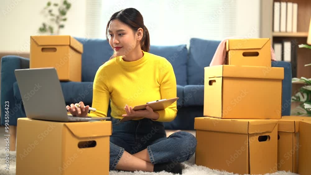 Young entrepreneur checks a list while working on a laptop, surrounded by cardboard boxes in a cozy living room, managing orders for her online store with a smile Young entrepreneur checks a list while working on a laptop, surrounded by cardboard boxes in a cozy living room, managing orders for her online store with a smile