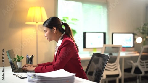 Businesswoman sits at her desk, working intently on her laptop. As time passes, fatigue sets in, causing her to slouch and hunch over while continuing to type