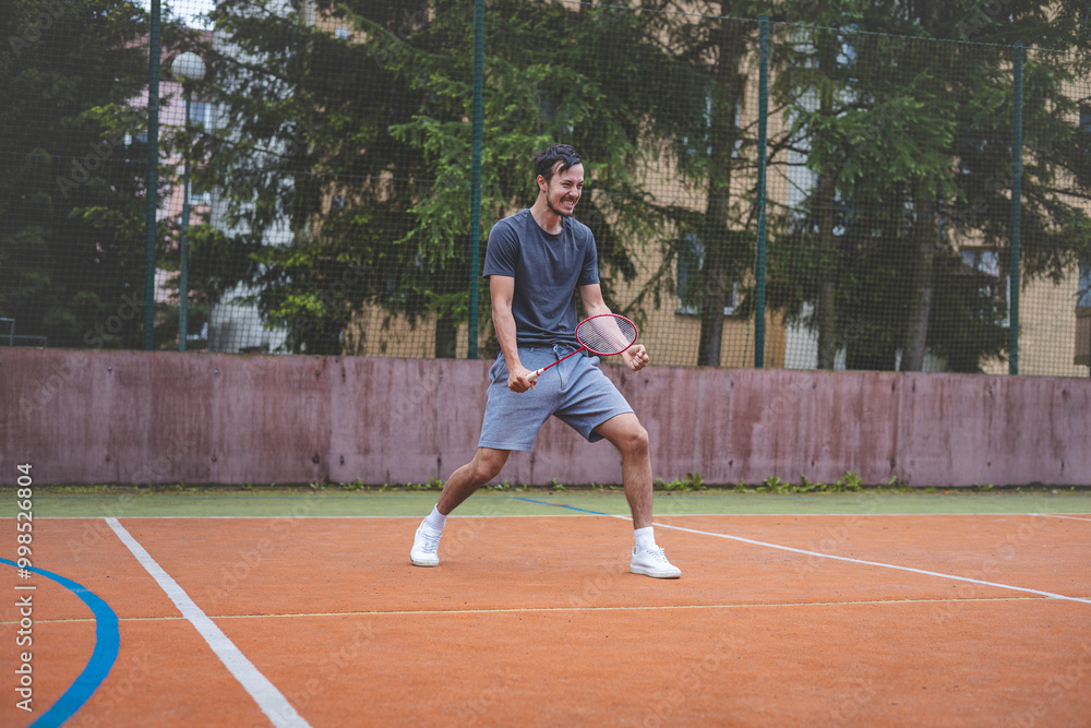 Badminton player celebrates a moment on an outdoor court, holding his racket in an energetic stance. His expression shows excitement and triumph, set against a backdrop of trees and buildings Badminton player celebrates a moment on an outdoor court, holding his racket in an energetic stance. His expression shows excitement and triumph, set against a backdrop of trees and buildings