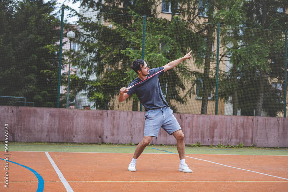 Badminton player celebrates a moment on an outdoor court, holding his racket in an energetic stance. His expression shows excitement and triumph, set against a backdrop of trees and buildings Badminton player celebrates a moment on an outdoor court, holding his racket in an energetic stance. His expression shows excitement and triumph, set against a backdrop of trees and buildings