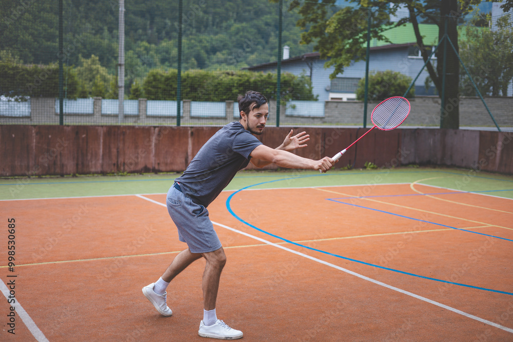 Badminton player focuses on the shuttlecock mid-air during a match on an outdoor court. With his racket raised, he prepares for the next shot, surrounded by an urban and scenic background Badminton player focuses on the shuttlecock mid-air during a match on an outdoor court. With his racket raised, he prepares for the next shot, surrounded by an urban and scenic background
