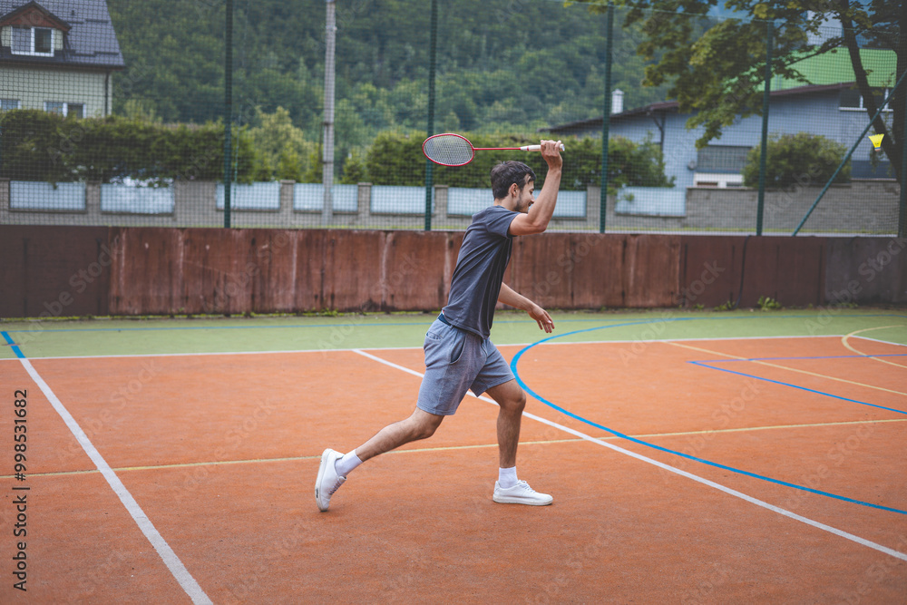 Badminton player focuses on the shuttlecock mid-air during a match on an outdoor court. With his racket raised, he prepares for the next shot, surrounded by an urban and scenic background Badminton player focuses on the shuttlecock mid-air during a match on an outdoor court. With his racket raised, he prepares for the next shot, surrounded by an urban and scenic background