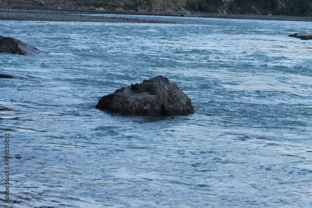 Obraz premium A striking black rock emerges from the clear blue waters of a flowing river, creating a captivating contrast against the vibrant blues. The gentle current surrounds the rock.