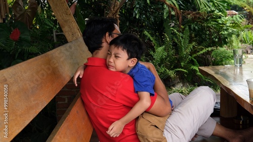 A father comforts his crying son in a heartfelt embrace while sitting outdoors. The boy seeks solace in his father’s arms, capturing a tender moment of love, care, and emotional support in nature.