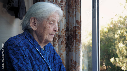Portrait of sad old woman looks hopefully out window at street through glass. Worrying or missing family relatives. Alone people in old age in nursing homes. Suffers nervous tics and tremor of hands